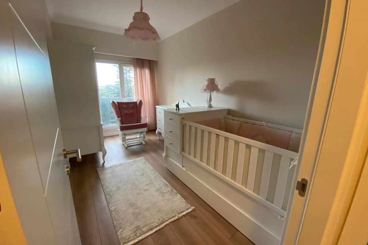 baby-bedroom-with-white-cradle-beige-carpet-on-wood-effect-tiles-pink-armchair-and-curtains-on-floor-to-ceiling-window-walls